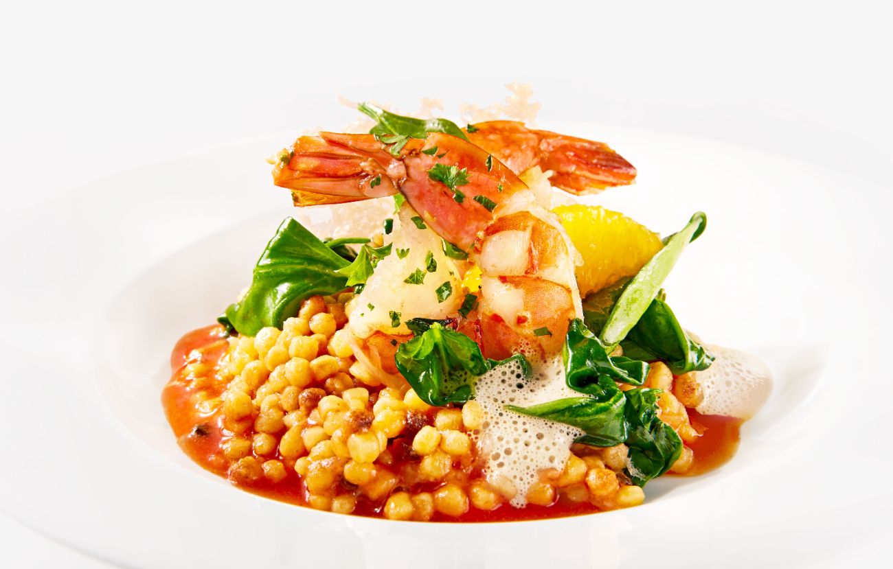 Der Zwischengang „Risotto“ von Fregola sarda mit Spinat, Orange und sautierten Garnelen in Krustentierschaum im PALAZZO Berlin.<br/>
