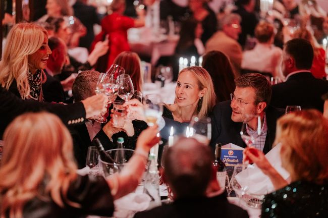 Die Gäste im PALAZZO Spiegelpalast sitzen an den Dinnertischen und prosten sich mit Weingläsern zu.