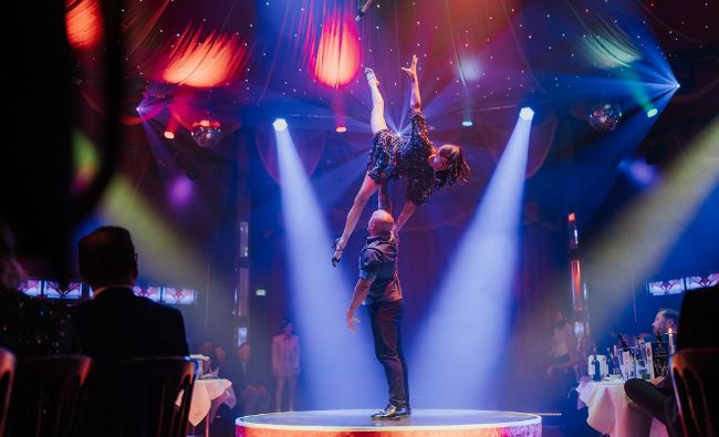 Olavo hält seine Partnerin Taylor mit einem Arm über seinem Kopf während sie einen Spagat macht.